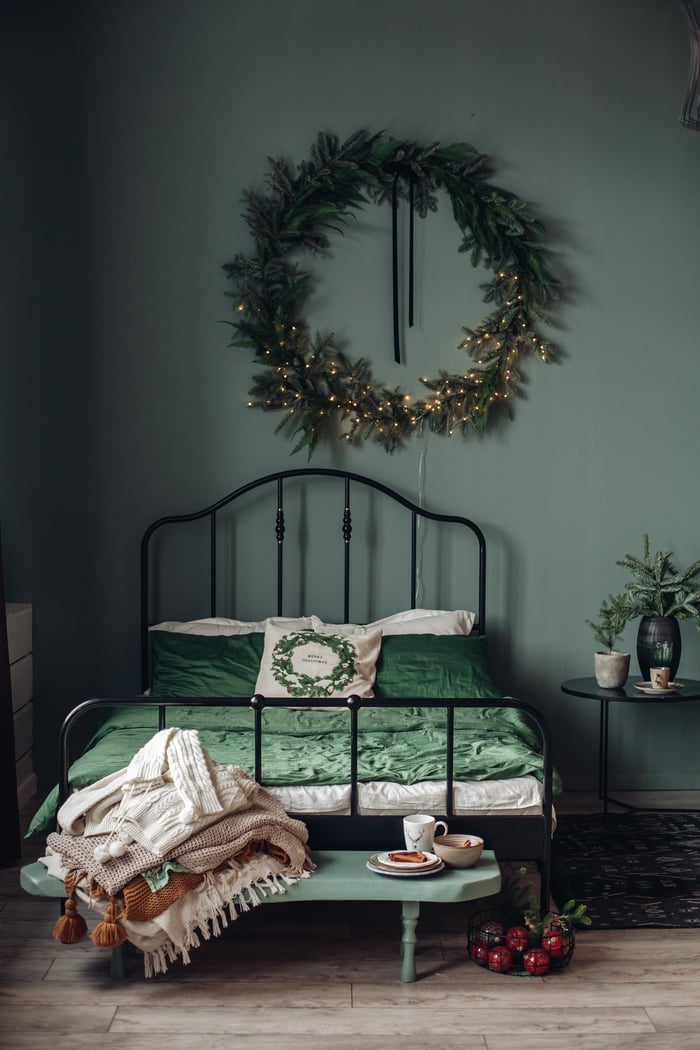 View over green modern room decorated for christmas holidays. Decorated and illuminated wreath on wall over bed, pine branches in vase on nightstand, and ornaments in wire basket on floor.
