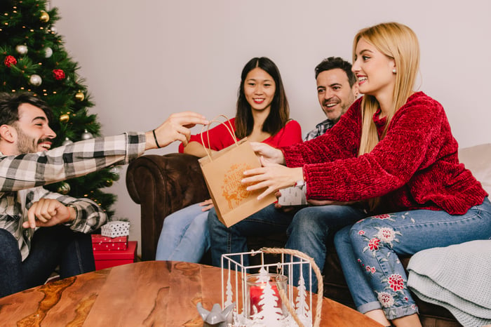 Group of four visible friends sitting in a living room during a holiday party. One friend chooses to steal the the present of a friend sitting across from them during a white elephant gift exchange