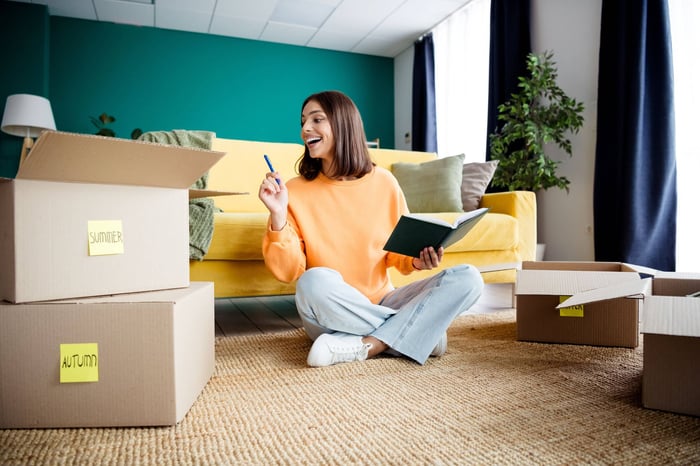 Young brunette woman in a cozy home living room with moving boxes writing in a notebook and planning a fresh start after a move to the best state to live in