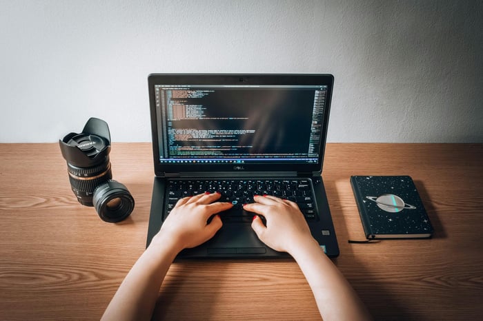 A woman coding on a laptop with a camera lens and notebook on a wooden desk in a home office.