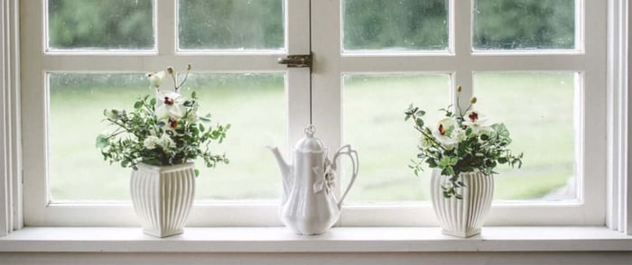 Older white windows in a fixer-upper home overlooking a blurred out backyard, decorated with flowers in vases and a tall, white tea pot.